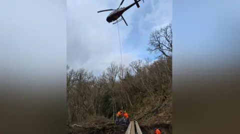 Westmorland and Furness Council View looking up at a helicopter with a dangling cable which has just been used to deposit three long pieces of timber - part of a bridge - at a woodland site. Two workmen in high visibility jackets are holding the end of the cable, with another one next to the timber. Behind them is a bank containing leafless trees.