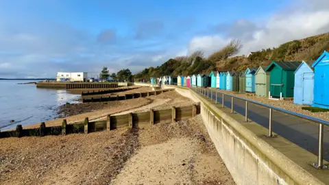 A delightful sunny winter's day at a beach. Beach huts run down the side of a pavement on the right-hand side looking out on to the sea and a stony beach. The sky is mainly blue with a few fair weather clouds.