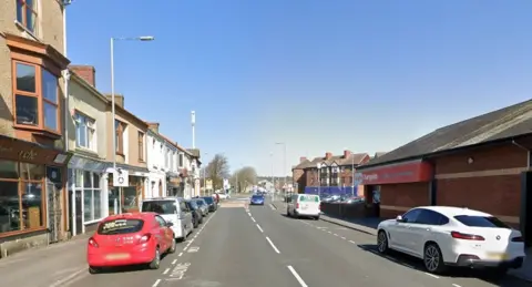 Google A road with cars parked on either side of it. There is a Home Bargains store just visible on the right side of the image with other shops on the left.