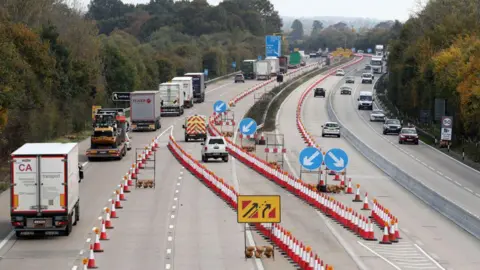 A picture of a motorway with many trucks on the left side. There is a row of traffic cones with two more cars merging into the left lanes. 