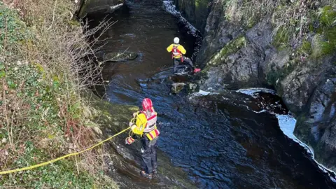 Cave Rescue Organisation Two cave rescue volunteers, wearing red hard hats, fluorescent yellow jackets and black waterproof trousers stand in a river, attached to yellow ropes. The riverbed has steep, rocky sides and grassy ledges.