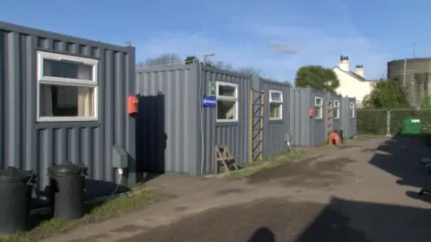BBC Three corrugated iron crates with windows in front of a row of houses.