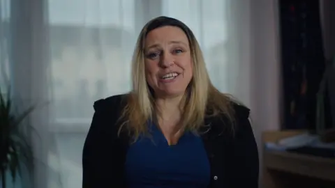 BBC/Alleycats TV Hillery wears a blue top and black jacket. She has blonde hair.