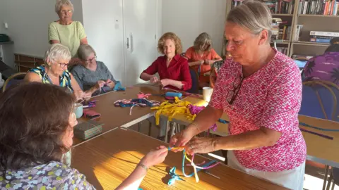A woman in a red and white top ties ribbons with the help of another woman, watched by other members of a sewing class 