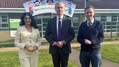 Shabina, a woman in a yellow suit, standing next to Jonathon, a man in a grey suit and Andrew Pakes, a man in a blue jumper and jeans, smiling for camera. 