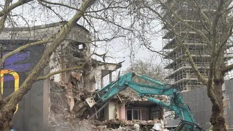 A green crane tears into a half-destroyed large building. Two trees are in the foreground and a tower with scaffolding is in the distance.