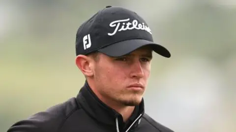 Tyler Weaver during a golf competition. He wears a black cap with the brand name Titleist on it. He wears a black jumper. He looks away from the camera.