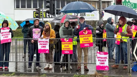 The picket line which has 12 teachers standing behind a meatl gate holding signs which read: OFFICIAL PICKET LINE and SUPPORT OUR STRIKE and they are branded by UCU. It is raining so the teachers are wearing anoraks and have umbrellas. Behind them is a Truro and Penwith College building. 