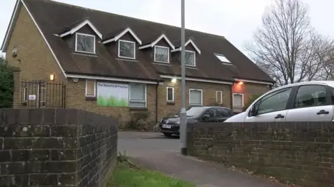 A general view of the Waterfield practice in Bracknell, a two storey building with a sign saying "The Waterfield Practice" and two cars parked in his parking lot.