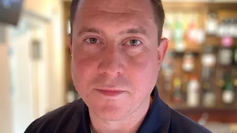 A man stood inside a pub looks into the camera.