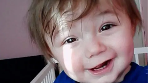 Family Photo A young boy with brown hair which is sweeping over his forehead. He is wearing blue top and is in a white cot as he smiles at the camera, his front baby teeth showing.