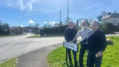 Three men stood behind a white road sign reading Pleasley Road with a road junction to the right of the image