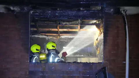Eddie Mitchell A missing window in a brick building, with black marks around the outside. Two firefighters in safety gear are hosing down a burned interior.