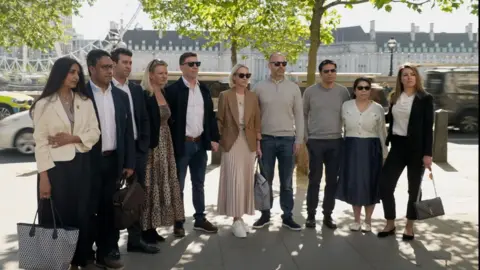 Ten people standing outside side by side in a row, looking away from the camera, with the river Thames in the background. Five of them are male, while the rest are women. Everyone is dressed in smart attire and most people are wearing sunglasses.