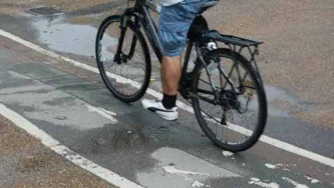 A man cycling on a cycle path. Only the lower half of his body is visible. He is wearing blue shorts, black socks and white trainers.