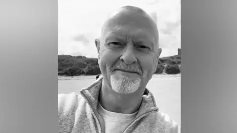 A black-and-white picture of Ian Pascoe, who is bald and has a goatee. The beach is in the background.