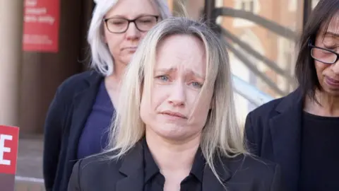 A woman with blonde hair wearing all black. She looks devastated and is holding back tears. There are people standing behind her also looking very upset.