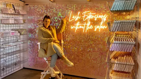 Lucy sitting on a tall white chair in her shop in front of a bright neon sign which says 'lash extensions without the salon'. The sign is on a glittery wall. There are shop displays either side of her with her products inside