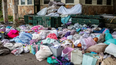 
Furniture and various colorful bags of clothes and other items sit on the dirt along with wet mattresses in the background 