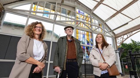 Cate Watkinson in a beige cardigan is standing next to Mike Davis wearing a flatcap and Rita Griskonyte in a beige jacket. They are all in front of the glass artwork