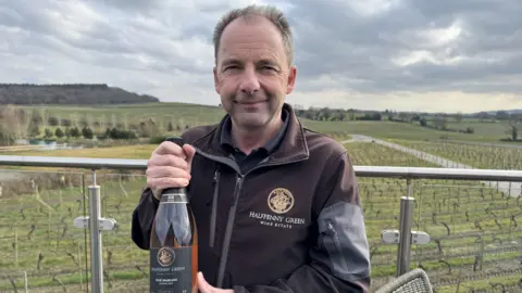 BBC A man with grey hair is wearing a brown and grey jacker with a circular gold logo on the breast. white writing on it says "halfpenny green wine estate". He is holding a bottle of rosé wine. He is on a balcony and beyond him is a vineyard with green grass and vines growing