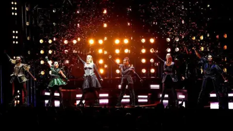 Getty Images A photo of a performance of Six The Musical. Six actresses are on a darkened stage, representing the six wives of Henry VIII.