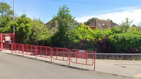 Google A pavement goes across the bottom of the image, with a red fence going across. Behind a row of green bushes, you can see the primary school.