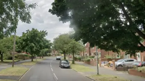 A suburban road lined with trees and houses. Parked cars, lamp-posts and other street furniture can be seen.