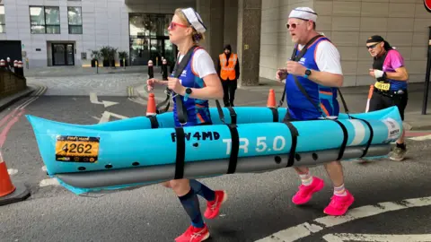 Nicky Ford/BBC Two runners wearing sailor hats navigate the race course while carrying a large, blue inflatable boat prop around their waists.
