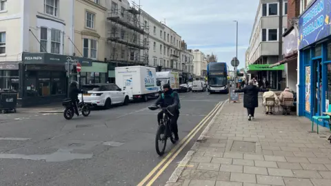 George Carden/BBC Western Road lined with shops and cafes. Cars and vans are parked on double yellow lines. Cyclists, cars and a bus are on the road. People sit outside on cafe chairs.
