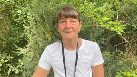 Teresa Atkins standing in front of trees at Ostler's Plantation. She is wearing a white t-shirt and has a lanyard around her neck. She has blue eyes and brown hair.