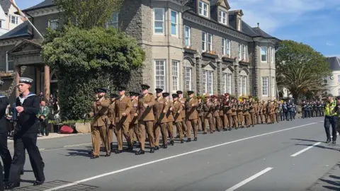 A military parade. A group of soldiers in tan uniforms march down the centre of the road past trees and an old building. 