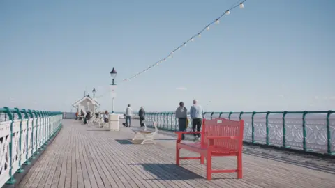 Bristish Heart Foundation A red bench on a traditional pier with wooden floorplanks and Victorian-era lampposts, railings and sheltered seating area at the far end.