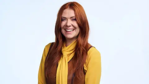 Sophie is wearing a yellow top with a brown waistcoat over the top. She has long red hair.