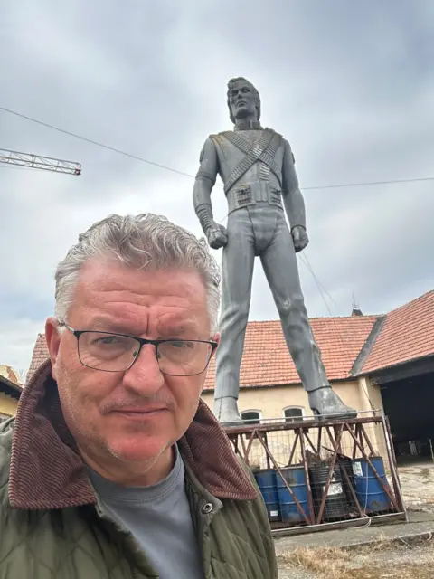 Franz Josef Zika A man with glasses and silver hair is taking a selfie in front of a Michael Jackson statue perched on a base made of oil drums. Parts of the statue look brown and stained white.