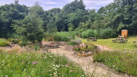 A view of the garden now, it has a path to a focal point which has a wired three legs of man in it, flower beds are overgrown, there are benches on a grassy area in the space.