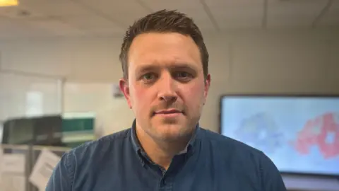 Alex Houston wears a navy, open collared shirt. He has short brown hair. Behind him is a white wall with a blurred TV screen with two maps of Northern Ireland. 