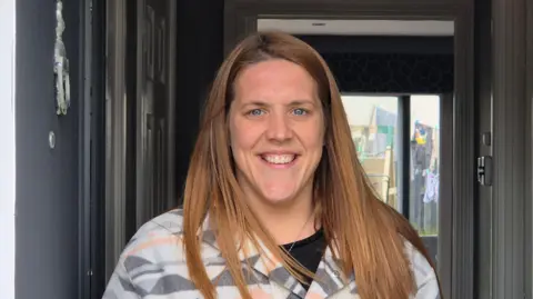 Karena Quaile - a woman with red hair past her shoulders, smiling into the camera while standing in the hall of her house
