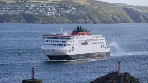 The Manxman ferry, which has red, white and blue livery, sailing into Douglas Harbour. The vessel is surrounded by blue sea, and in the background white houses can be seen on a green headland.