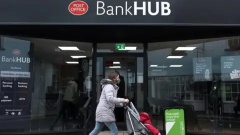 Getty Images A woman wearing a mask is pushing a child in a buggy past a banking hub which also has the red Post Office sign. 