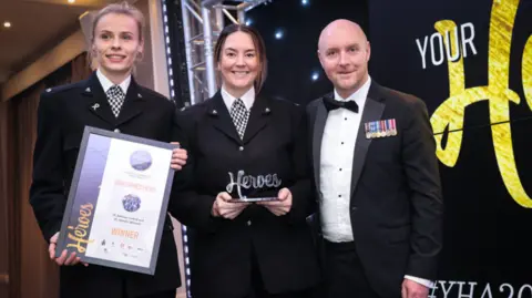 Two women in formal police uniform stand alongside a man with medals on his dinner jacket. One of the offices is holding a certificate, the other a trophy spelling out the word Heroes