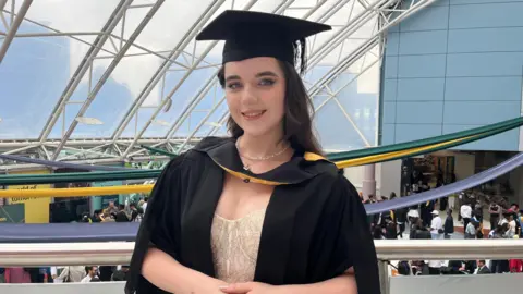 A woman with long dark hair is wearing a graduation gown and cap smiling