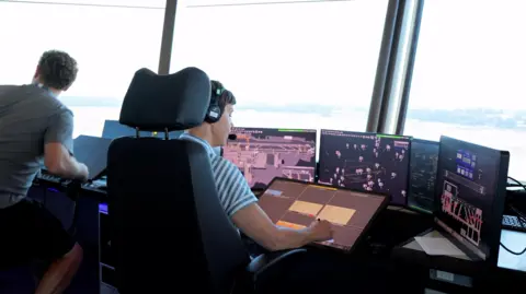 An interior picture in the air traffic control room at Gatwick Airport. Controllers sit in front of several screens and have a view over the tarmac.