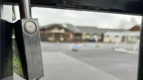 BBC An extreme close up of a silver padlock attached to black gates. A school is out of focus in the background.