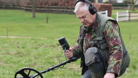 David Stuckey is crouched on one knee in a field. He is holding a metal detector and looking at a small monitor. He is wearing a camouflage jacket and has knee pads over his blue trousers, which are tucked into green boots. he has short greying hair and is wearing headphones on his head