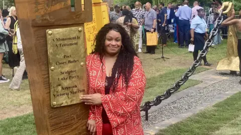 A young black woman with dark curly hair points at the gold plated sign on the Windrush monument 'A Monument is a Testament', which shows her name. She is wearing a red kimono style top with a white pattern on it 