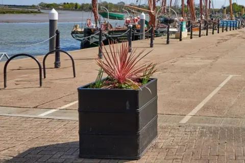 MALDON DISTRICT COUNCIL Positioned on a promenade, a black container with plants growing out of it.
In the background there are bollards and a place to lock bicycles. The river and boats can also be seen.