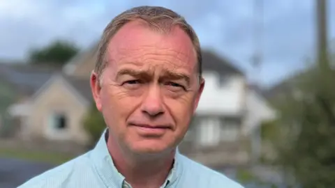 A close-up headshot of Liberal Democrat MP Tim Farron, wearing a light blue collared shirt. He has sandy short hair and is wearing a serious expression.