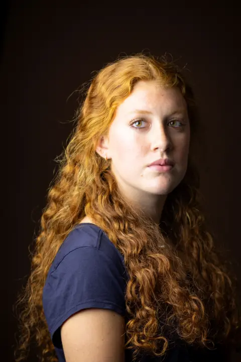 Kieran Dodds Close-up studio portrait of a person with long, curly red hair cascading over the shoulders, wearing a dark short-sleeved top. The person is turned slightly to one side while looking toward the camera. Soft, warm lighting highlights the texture of the hair against a dark brown background.