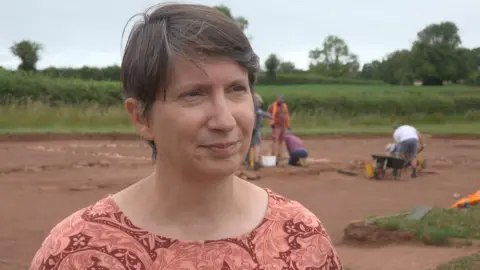 Dr Susan Greaney is stood in front of people digging at the soil. She has short hair and is wearing a pink top with leaves on it.